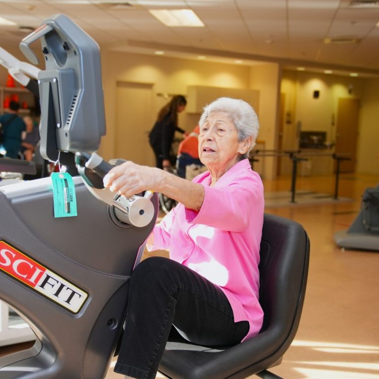 Elderly woman with white hair exercising on a SCIFIT recumbent bike in a rehabilitation gym.