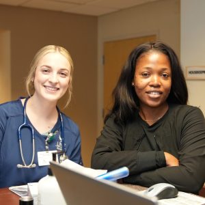 Two smiling women, one a blonde nurse in scrubs and the other in a black jacket, sitting at a desk.