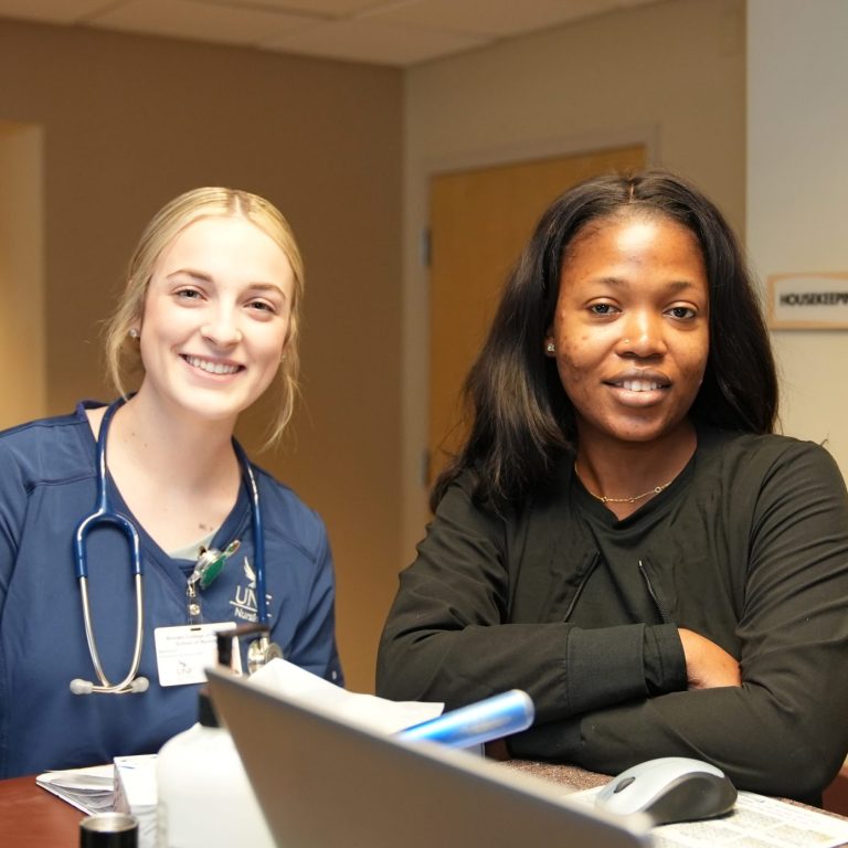 Two smiling women, one a blonde nurse in scrubs and the other in a black jacket, sitting at a desk.