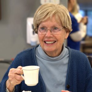 A smiling elderly woman with short blonde hair and glasses, wearing a blue cardigan and turtleneck, holding a white cup.