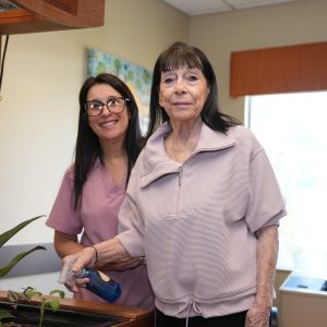 A senior woman waters plants with a spray bottle, assisted by a smiling caregiver.