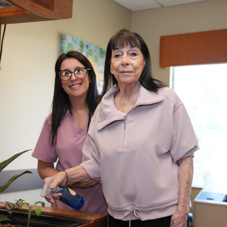 A senior woman waters plants with a spray bottle, assisted by a smiling caregiver.