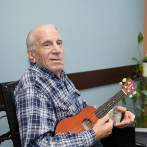 Senior man in a blue and white plaid shirt, seated in a wheelchair, holding a ukulele with a bandaged hand, looking at the...