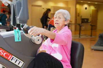 Elderly woman with white hair exercising on a SCIFIT recumbent bike in a rehabilitation gym.