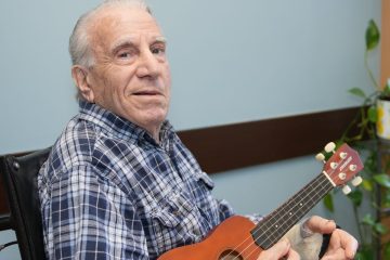 Senior man in a blue and white plaid shirt, seated in a wheelchair, holding a ukulele with a bandaged hand, looking at the...