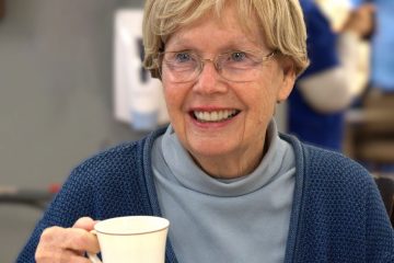 A smiling elderly woman with short blonde hair and glasses, wearing a blue cardigan and turtleneck, holding a white cup.