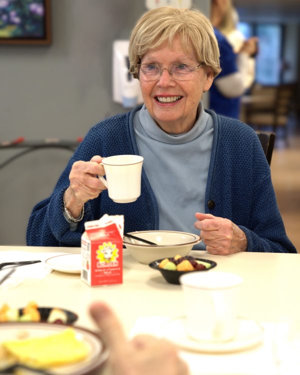 Elderly woman with blonde hair and glasses smiles while holding a mug at a dining table with breakfast items.